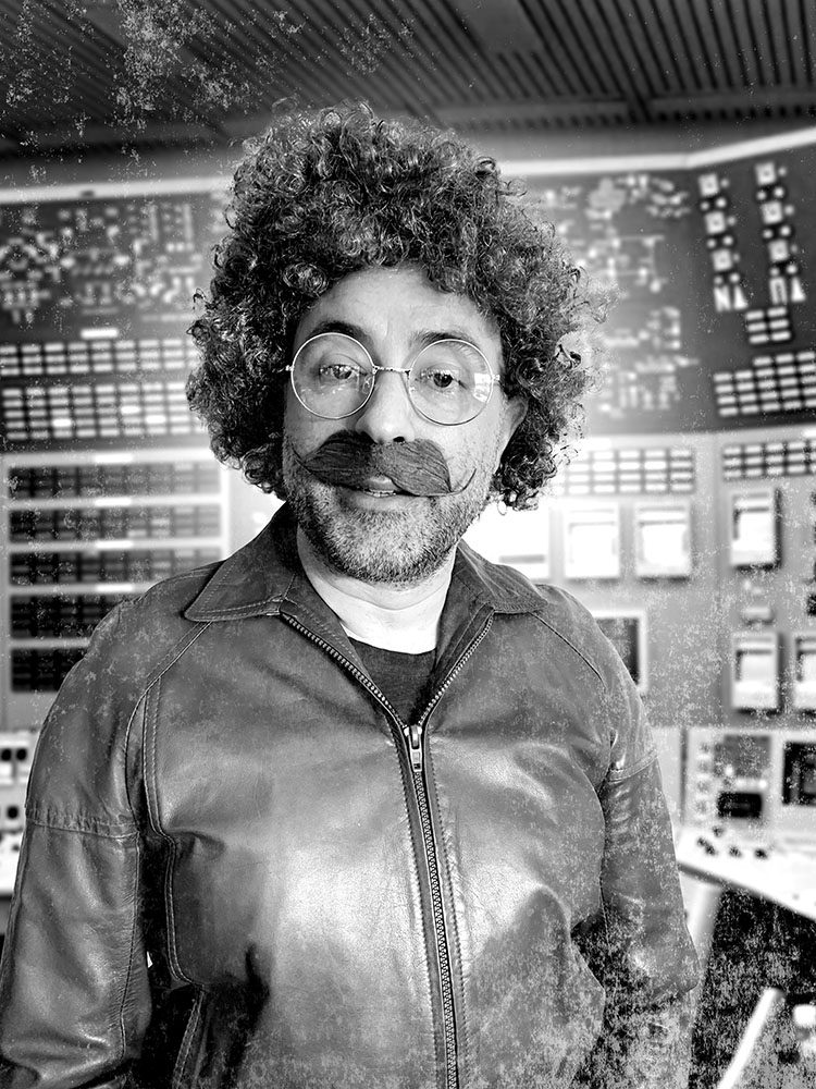 A man with curly hair, glasses, and a mustache wearing a leather jacket, standing in front of a complex control panel with various knobs and screens.